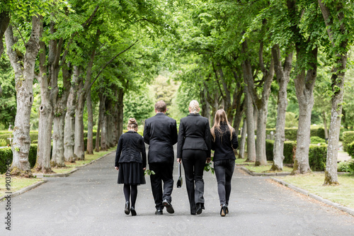 Familie läuft auf Allee im Friedhof in Trauer mit Blumen in der Hand