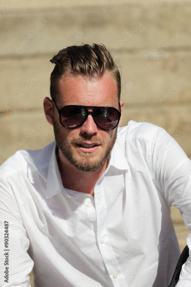 Portrait of a good looking man in his 20s, wearing a white shirt with dark sunglasses, sitting down outside on a set of steps on a sunny summer day. Portrait of a good looking man in his 20s, wearing a white shirt with dark sunglasses, sitting down outside on a set of steps on a sunny summer day.