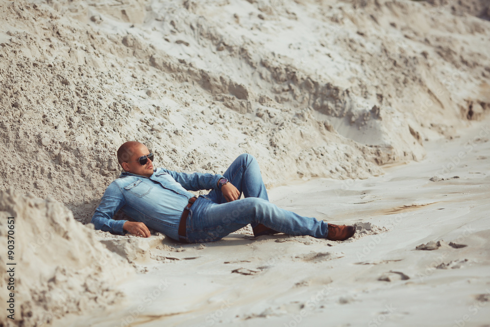 gorgeous young man in glasses lies in jeans clothes on the sand
