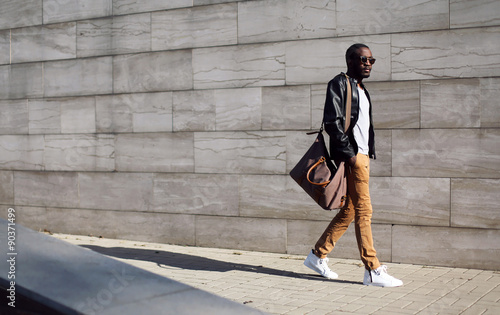Street fashion, stylish african man in black rock leather jacket with bag walking in evening city