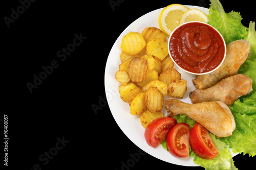 chicken legs on a white plate with slices of tomato and lettuce and french fries and ketchup top view isolated on black background