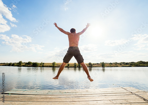 Happy man jumping on pier with lake and sky in background © Maksim Kostenko