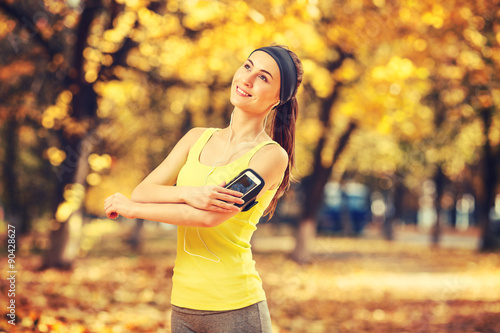 Fototapeta Naklejka Na Ścianę i Meble -  Running woman. Runner is training and listening to music. Female fitness model training outside in the sunny autumn park. Athlete resting in the fall outdoors and using smart phone. Sport lifestyle.