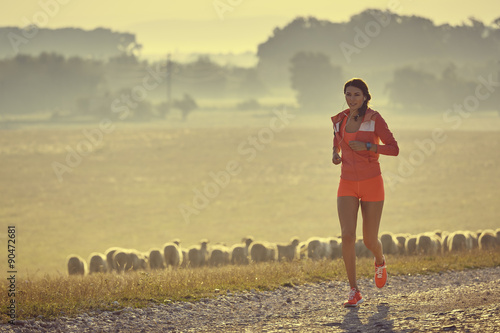 Beautiful fit young lady in red summer sportswear running, jogging on a country road while listening to music in earphones, early in the morning. Calories burning. Health care. Willingness concept.