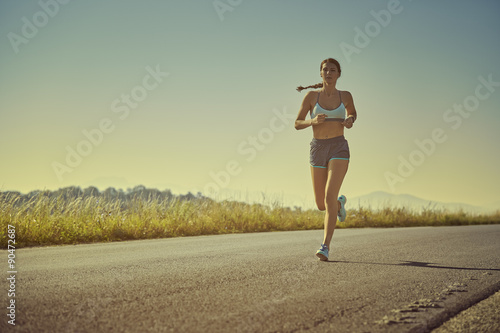 Active sporty woman in summer sportswear running, sprinting on a road at sunrise or sunset. Health care, body care, healthy lifestyle, willingness concept. Toned color edit. © Photosebia
