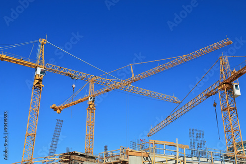 Three construction cranes and building against blue sky.