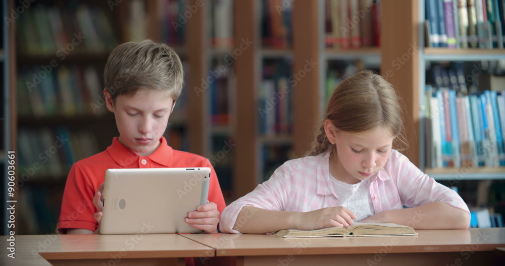 Two classmates sitting at desk: boy reading on tablet computer while girl reading paper book Two classmates sitting at desk: boy reading on tablet computer while girl reading paper book