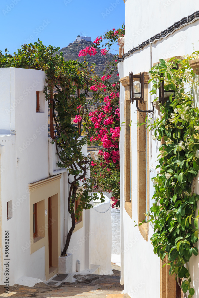 Narrow street in Chora on the island of Patmos, belonging to the Dodecanese, Greece. Beautiful flowers of bougainvillea contrast to the white walls of houses and blue sky