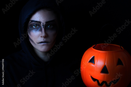 Girl dressed in scary face paint with pumpkin bucket on black ba