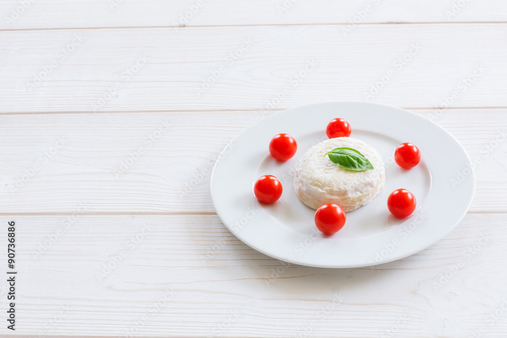 Camembert round entirely with cherry tomatoes and basil leaf on a white plate and a white wooden background Camembert round entirely with cherry tomatoes and basil leaf on a white plate and a white wooden background