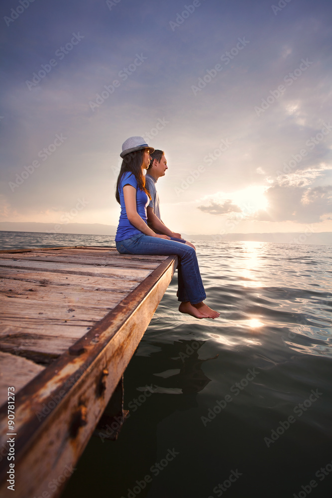 Fototapeta premium Side view of young couple sitting on the pier watching sunset or sunrise