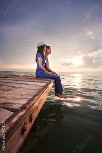 Side view of young couple sitting on the pier watching sunset or sunrise
