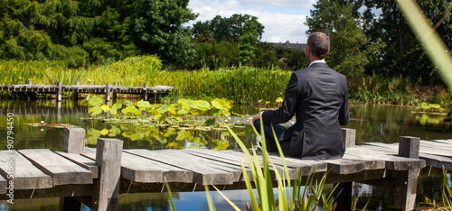 corporate green break - meditating middle age male professional sitting on a wooden bridge in the middle of an asian-like green pond for company wellbeing,back view with park foreground