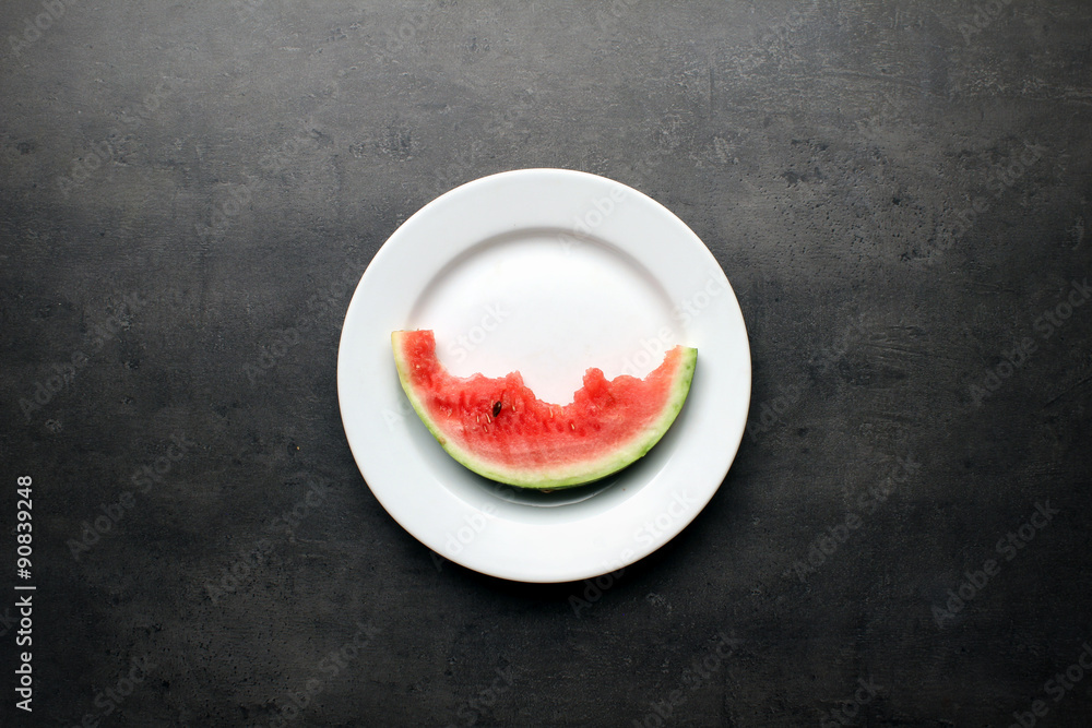 Watermelon slice with bites on white plate and dark kitchen table background Watermelon slice with bites on white plate and dark kitchen table background