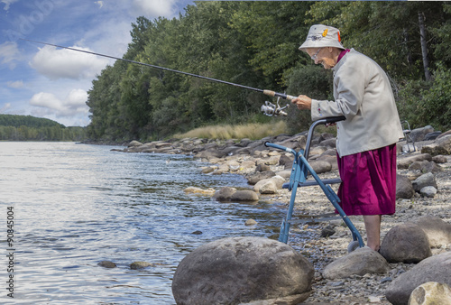 horizontal image of an elderly senior woman with a walker fishing at the edge of the lake with trees and rocks lining the shoreline in the summer time
