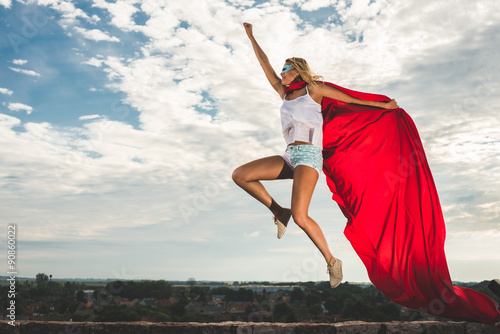 Blonde woman in red dress and red mantle jumping outdoor as a superhero against blue sky © Laszlo