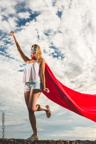 Blonde woman in red dress and red mantle posing outdoor as a superhero against blue sky