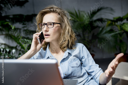 Beautiful young businesswoman using mobile phone while working with laptop in office. Upset or dissapointed expression at her face