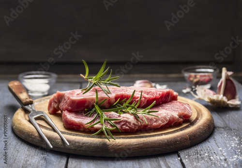 raw meat with rosemary, garlic, salt and pepper on a wooden board with a for, top view