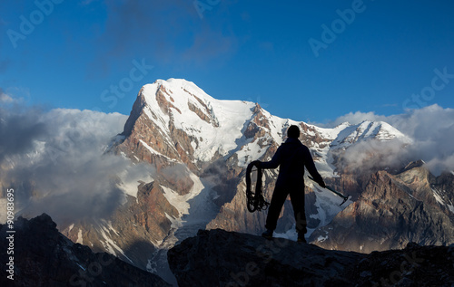 Alpine Climber Arranging Descent with Rope and Ice Axe Silhouette Man Staying on Top of Rock Cliff Holding Climbing Gear Stormy Clouds and Peaks Illuminated bright Morning Sun