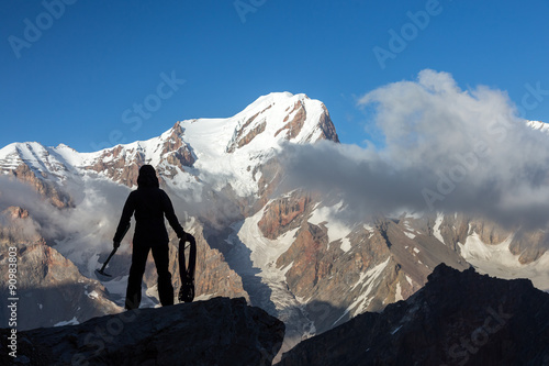 Alpine Climber Arranging Descent with Rope and Ice Axe Silhouette Woman Staying on Top of Rock Cliff Holding Climbing Gear Stormy Clouds and Peaks Illuminated bright Morning Sun