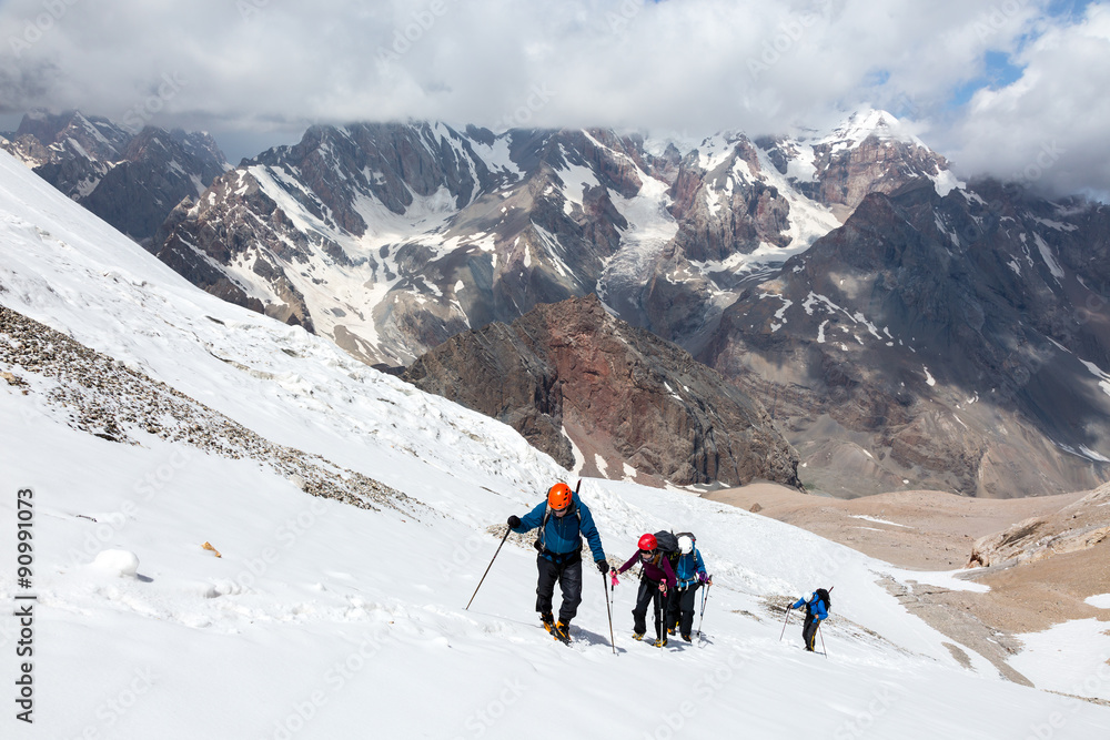 Group of Hikers Walking on Snow and Ice Terrain Large Group of People Sport Clothing Going Heavy Load Backpacks Climbing Gear Up Mountain Peaks Sunlight Cloud Sky Majestic Summits Background