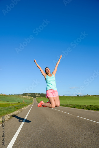 Happy woman jumping for celebrating running and sport success. Female athlete raising arms to the sky during training on countryside road.