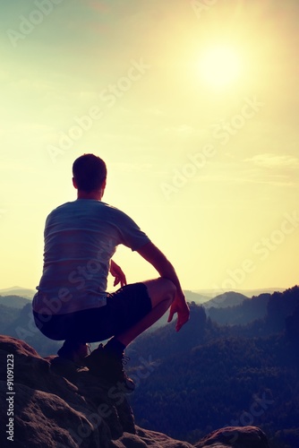 Young man in black sports pants and grey shirt is sitting on cliff's edge and looking to misty valley bellow