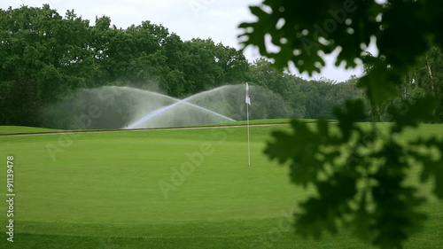 in the front is a blurred branch of an oak and in the background the golf course with sprinklers and a white golf flag.