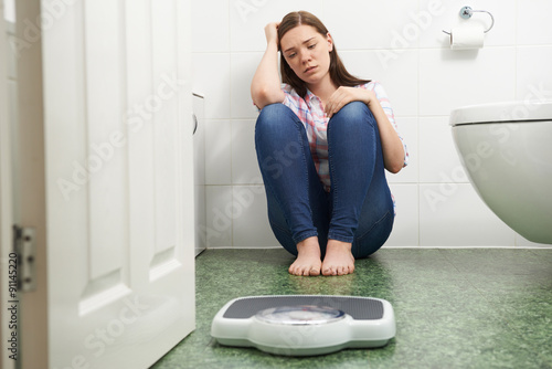 Unhappy Teenage Girl Sitting On Floor Looking At Bathroom Scales