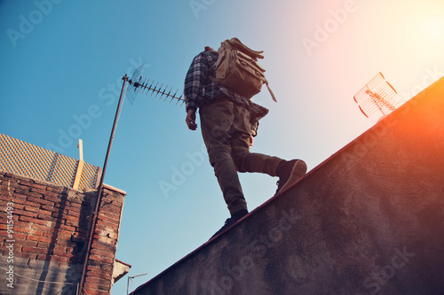 Billede på lærred Brave man with backpack walking over high wall on the edge, on the rooftop (inte