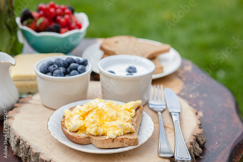 Fototapeta Naklejka Na Ścianę i Meble -  Breakfast in the garden: scrambled eggs on toast and yogurt with berries on the dark wooden table, selective focus