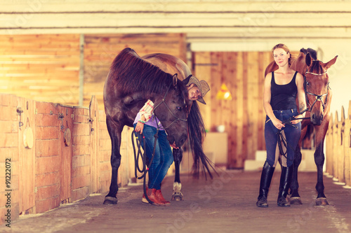 Cowgirl and young woman in stable with horses.