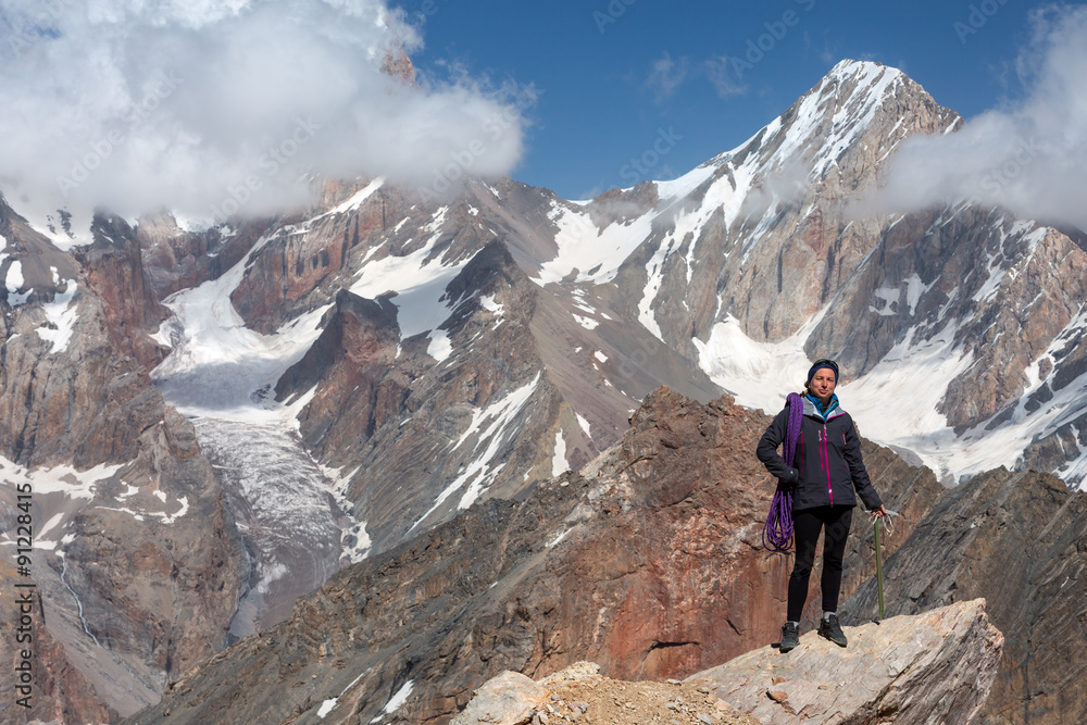 Fototapeta premium Female Climber Holding Ice Axe and Alpine Rope Full Body Portrait of Smiling Woman Staying on Pointed Rock Top Sporty Clothing High Altitude Mountain Landscape Blue Sky
