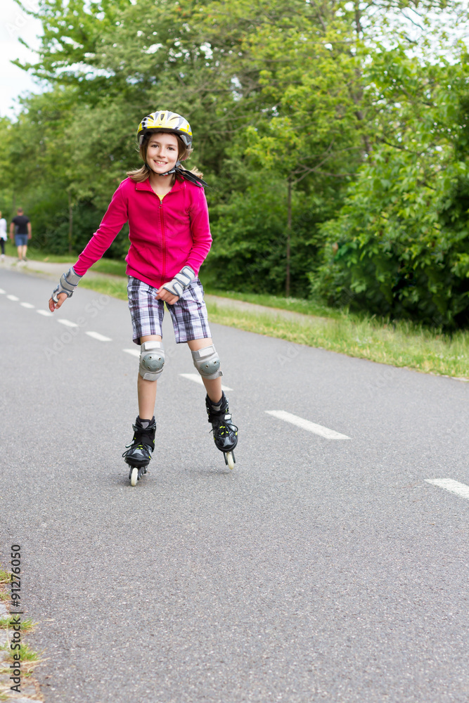 Obraz premium Smiling little girl roller skating in a public park. The girl has a helmet and protective knee and wrist pads