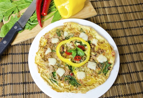 A spinach, chili, mint and pepper omelette in a dish over bamboo table cloth and chopping board with knife and raw vegetables on while background. A healthy diet meal omelet. photo