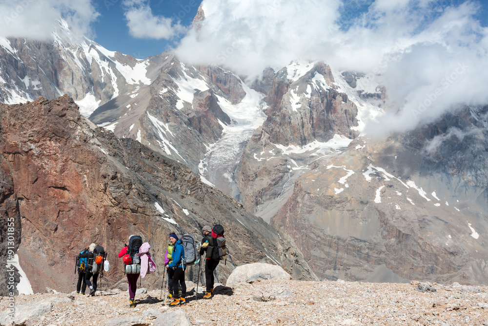 Fototapeta premium Backpackers Team Walking on Rocky Trail Group of People in Sporty Style Clothing with Backpacks and Walking Poles Mountain Landscape with Blue Sky and Clouds on Background