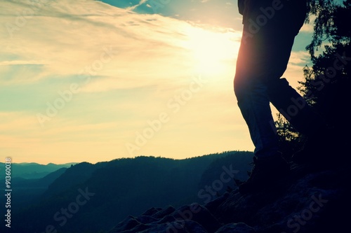 Woman hiker legs in tourist boots stand on mountain rocky peak. Sun n background