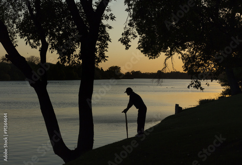 horizontal silhouette of an old man leaning on walking cane looking out across the lake at sunset with trees lining the banks on a summer evening.