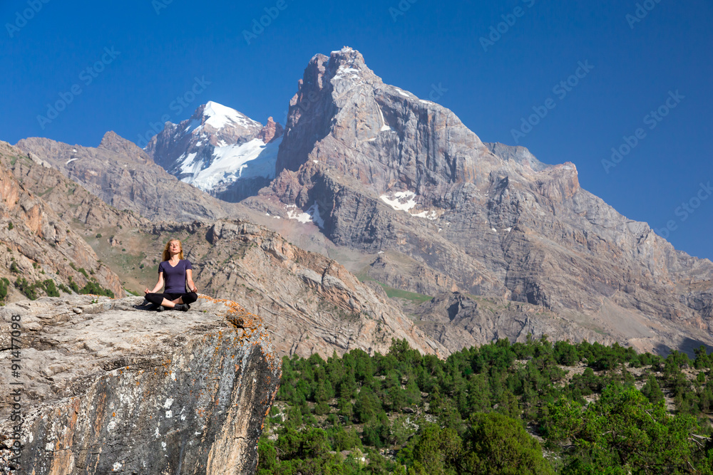 Young woman doing exercise Female Athlete Sitting in Yoga Zen Mediation Pose Mountain Panoramic Landscape Outdoor Orange Rocks Green Forest Clear Blue Sky High Summit with Snow Top on Background