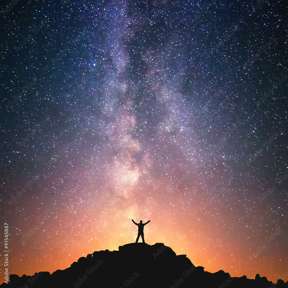 Man and the Universe. A person is standing on the top of the hill next to the Milky Way galaxy with his hands raised to the air. - obrazy, fototapety, plakaty 