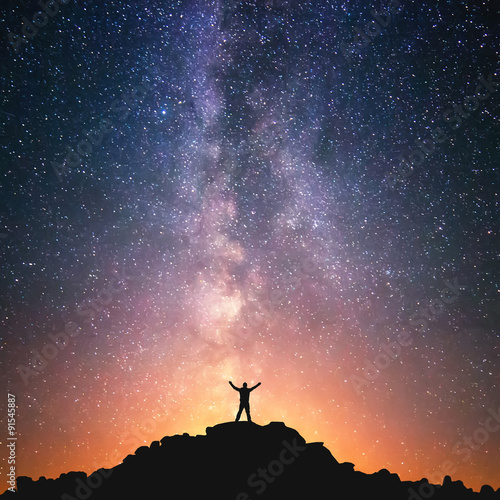 Man and the Universe. A person is standing on the top of the hill next to the Milky Way galaxy with his hands raised to the air.