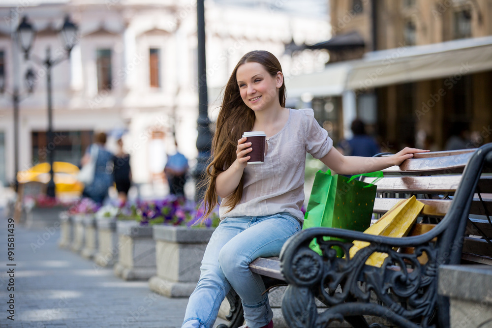 Young beautiful woman sitting on a bench on the street with coffee cup in hand and shopping bags. Drinking coffee and smiling.
