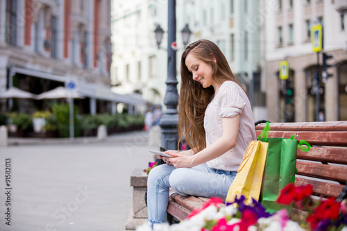 Young beautiful woman sitting on a bench with digital tablet next to the shopping bags. Relaxing and using the device with happy smile
