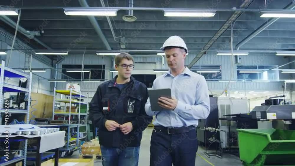 Engineer in Hard Hat and Factory Worker are Walking through Production Facility Engineer in Hard Hat and Factory Worker are Walking through Production Facility