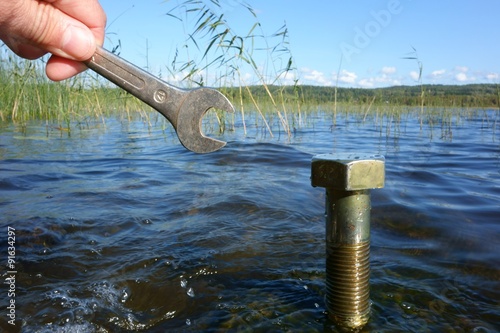 Male hand holding a wrench in front of a large bolt coming out of the lake water on a sunny Autumn afternoon in Finland with ecology, environmental protection and green concept idea. photo