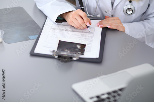 Close up of female doctor filling up a medical history form, sitting at the table in hospital