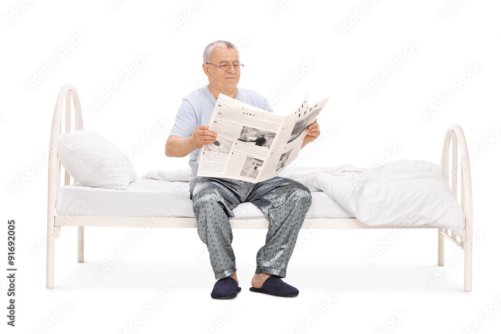 Senior in pajamas reading a newspaper seated on bed