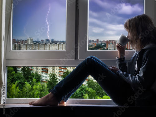 Девушка сидит на подоконнике, пьет из чашки и смотрит в окно. За окном молния, гроза, город. Непогода, домашний уют, вкусный напиток - чай или кофе 