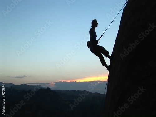 Long-shot of a rock climber silhouetted against a golden-hour-sky rappelling down a cliff. photo